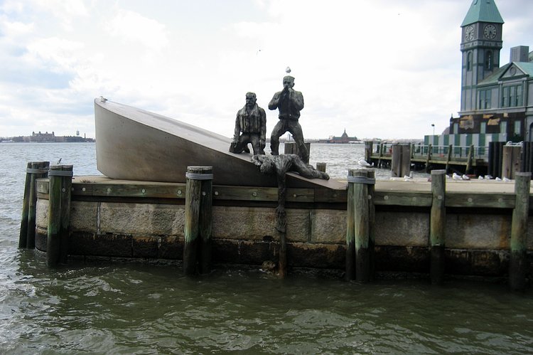 NYC - Battery Park: American Merchant Mariners Memorial | Commissioned by the American Merchant Mariners' Memorial, Inc., this memorial was conceived in 1976. In 1988, after an extensive competition, the artist Marisol Escobar (b. 1930), known as Marisol, was chosen to develop her design. Situated off-shore from the north end of Battery Park and just south of Pier A, the monument stands on a rebuilt stone breakwater in the harbor. The bronze figural group and boat are based on an actual historical event; during World War II, a Nazi U-boat attacked a merchant marine vessel, and while the marines clung to their sinking vessel, the Germans photographed their victims. Marisol developed a series of studio sketches from this photograph, then fashioned a clay maquette as her winning design proposal for the monument. The work was dedicated on October 8, 1991.