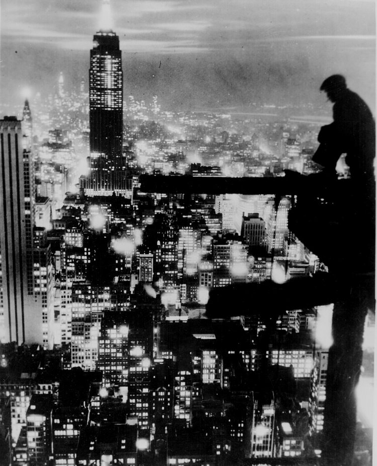 A man sits on a girder above New York City at night, ca. 1935
