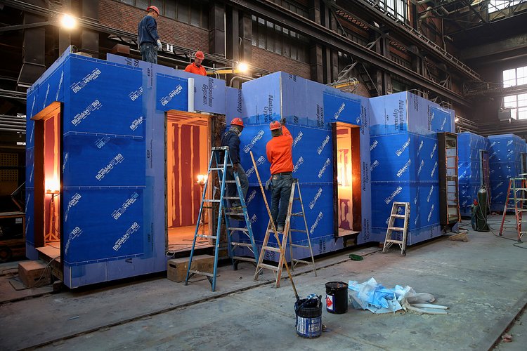 The zig-zag of the building facade is evident as workers apply a waterproof layer to a corner module.