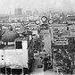 Luna Park on a busy weekend looking north along Jones Walk from entrance on Surf Ave. Women in dresses and men in suits with straw hats. Large industrial gas tanks loom in the distance. Coney Island. Brooklyn, New York.  1925