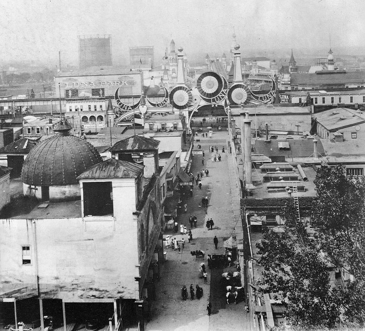Luna Park on a busy weekend looking north along Jones Walk from entrance on Surf Ave. Women in dresses and men in suits with straw hats. Large industrial gas tanks loom in the distance. Coney Island. Brooklyn, New York.  1925