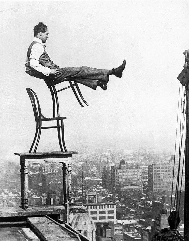 A daredevil balances on the back legs of a chair atop other furniture, 20 stories up in New York, circa 1920. 